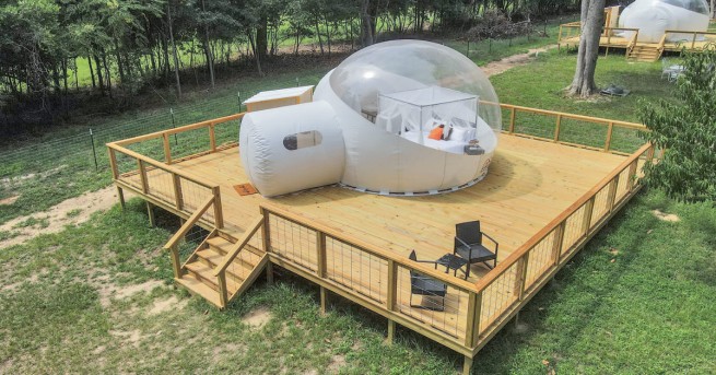 Transparent glamping bubble dome on a raised wooden deck with a bed inside, surrounded by trees and open green landscape.